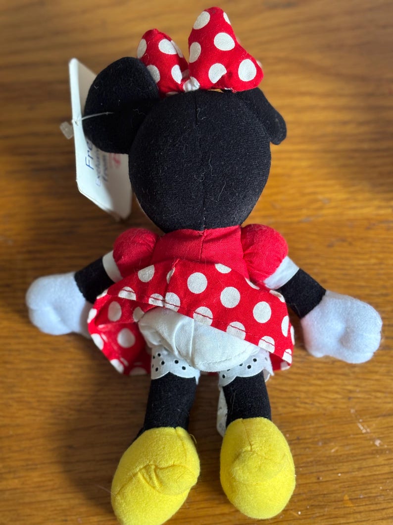 Puede incluir: Mu&ntilde;eca de peluche de Minnie Mouse, vista desde atr&aacute;s, con un vestido y un lazo rojos, ambos adornados con lunares blancos. La mu&ntilde;eca tiene orejas, brazos y piernas negros, con guantes blancos y zapatos amarillos. Una etiqueta est&aacute; adjunta.