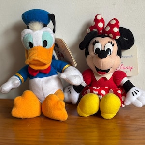 Puede incluir: Dos mu&ntilde;ecos de peluche de los personajes de Disney: Pato Donald y Minnie Mouse. El Pato Donald tiene un gorro de marinero azul, pico naranja y pajarita roja. Minnie Mouse lleva un vestido rojo de lunares y un lazo. Ambos tienen etiquetas.