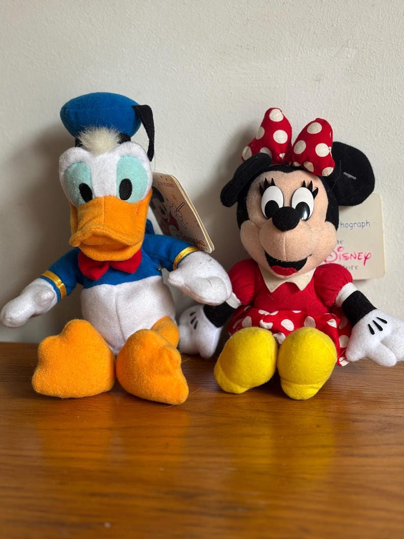 Puede incluir: Dos mu&ntilde;ecos de peluche de los personajes de Disney: Pato Donald y Minnie Mouse. El Pato Donald tiene un gorro de marinero azul, pico naranja y pajarita roja. Minnie Mouse lleva un vestido rojo de lunares y un lazo. Ambos tienen etiquetas.