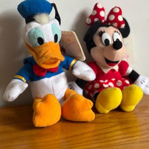 Puede incluir: Dos peluches de personajes de Disney: Pato Donald y Minnie Mouse. El Pato Donald lleva un traje de marinero azul, un sombrero negro y tiene un pico y pies naranjas. Minnie Mouse viste un vestido rojo con lunares blancos y un lazo a juego. Ambos tienen etiquetas.