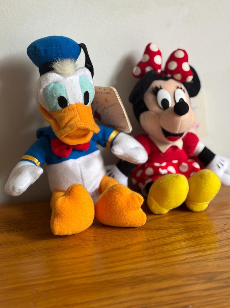 Puede incluir: Dos peluches de personajes de Disney: Pato Donald y Minnie Mouse. El Pato Donald lleva un traje de marinero azul, un sombrero negro y tiene un pico y pies naranjas. Minnie Mouse viste un vestido rojo con lunares blancos y un lazo a juego. Ambos tienen etiquetas.