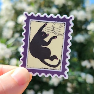 May include: A sticker in the shape of a postage stamp featuring a black cat in a playful pose. The cat is on a cream-colored background with a purple border. The stamp has a postmark with the number 13.