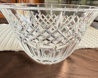 Puede incluir: Un cuenco de cristal tallado transparente con un patrón de diamantes. El cuenco tiene una forma ancha y acampanada y se asienta sobre una superficie marrón. El intrincado diseño capta la luz, creando un efecto brillante. Este cuenco decorativo es adecuado para servir o exhibir.