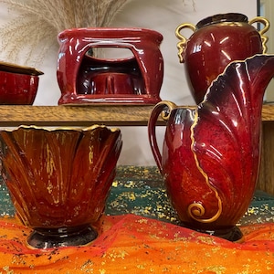 May include: A collection of decorative ceramic pieces in shades of red and gold. Includes a pitcher, a vase, a small box, and a candle holder. The pieces are displayed on a wooden shelf and a colorful fabric.