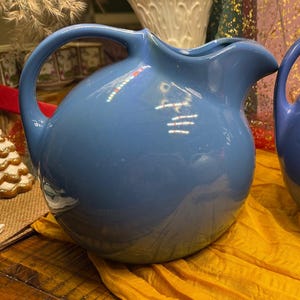 May include: Two ceramic pitchers in shades of blue. The pitchers have a rounded body, a curved handle, and a spout. They are sitting on a yellow cloth. The pitchers are likely for serving beverages.