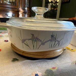 May include: A white casserole dish with a clear glass lid. The dish has a floral iris design in purple and green. It sits on a wooden trivet. A clear glass handle is on the side.