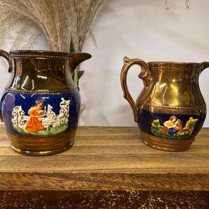 May include: Two antique ceramic pitchers with gold and brown finishes. One pitcher features a blue band with a painted scene of a person and animals. The other pitcher has a similar design.