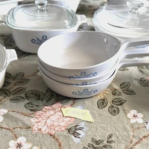 Op de afbeelding: Een verzameling witte CorningWare-schalen met blauwe bloemmotieven. De set bevat verschillende maten ovenschalen met glazen deksels, kommen en een kleine bak met een deksel. De schalen zijn gerangschikt op een tafelkleed met bloemenpatroon.