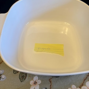 May include: A square white ceramic baking dish with rounded corners and integrated side handles. Inside the dish, a small yellow rectangular adhesive note is placed, featuring the handwritten text P-54B in black ink on a single lined section.
