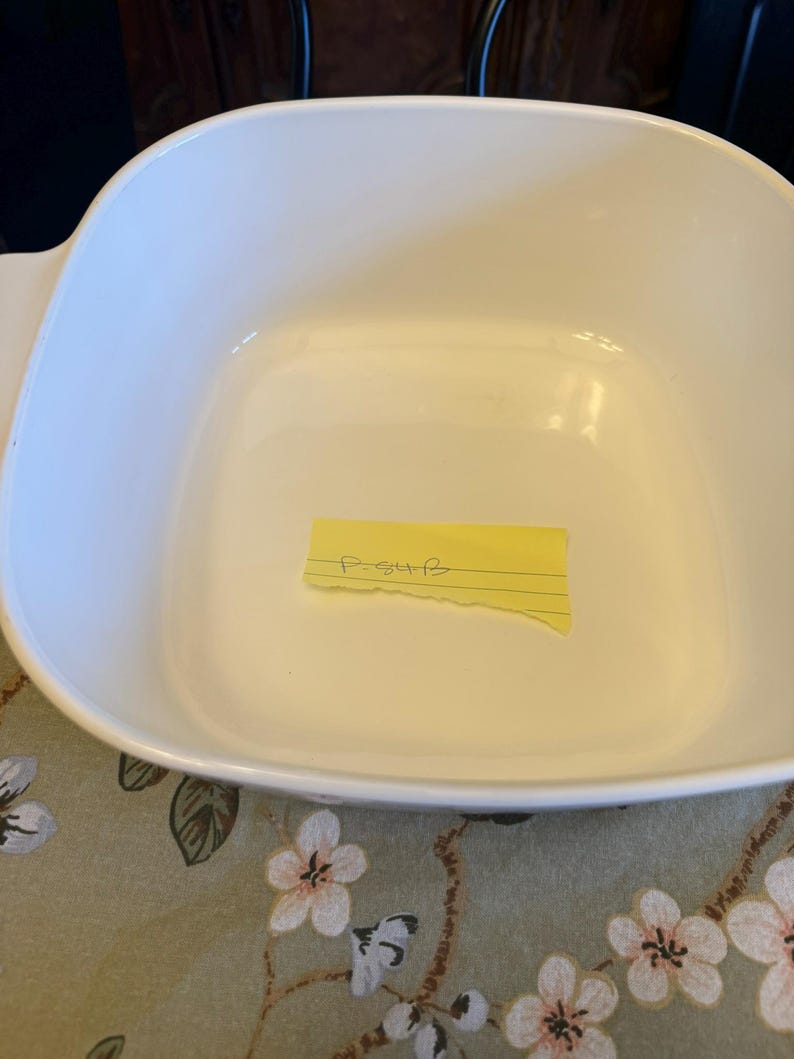 May include: A square white ceramic baking dish with rounded corners and integrated side handles. Inside the dish, a small yellow rectangular adhesive note is placed, featuring the handwritten text P-54B in black ink on a single lined section.