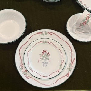 May include: A white ceramic dinnerware set featuring a swirling texture, a thin green rim, and a central motif of a green basket filled with greenery tied with a red ribbon. The set includes a bowl, a dinner plate, a salad plate, and a teacup.