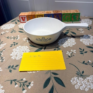 May include: A white ceramic saucepan with a long handle, featuring a blue floral pattern on the exterior. A yellow rectangular card with the handwritten text P-81-B is placed in front of the bowl, and wooden blocks spell out VINTAGE.