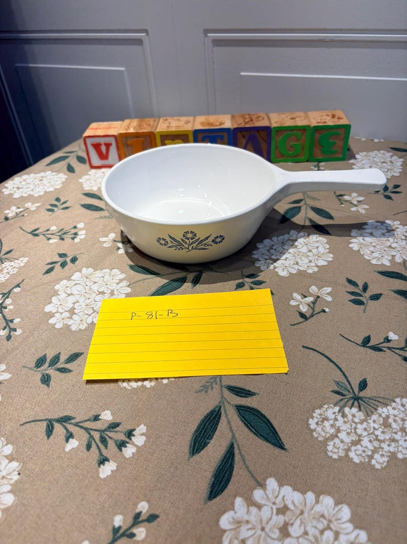 May include: A white ceramic saucepan with a long handle, featuring a blue floral pattern on the exterior. A yellow rectangular card with the handwritten text P-81-B is placed in front of the bowl, and wooden blocks spell out VINTAGE.