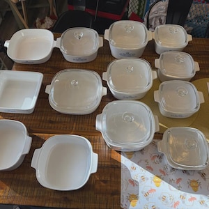 May include: A collection of white ceramic baking dishes and casserole dishes with glass lids. Some dishes have blue floral accents. The dishes are arranged on a wooden table, suggesting a kitchen or dining setting.
