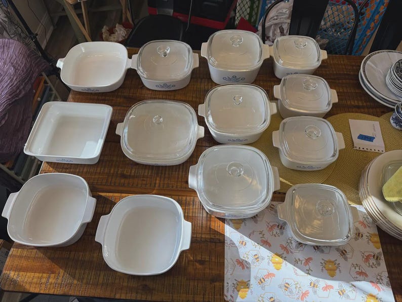 May include: A collection of white ceramic square and rectangular baking dishes, some with clear glass lids and integrated side handles. The dishes feature a subtle blue floral pattern on the exterior, arranged on a wooden surface with stacks of plates.