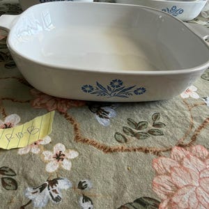 May include: A white ceramic square baking dish with rounded corners and integrated side handles. The exterior features a blue floral pattern consisting of stylized cornflowers and leaves centered on the side. The interior is smooth and white.