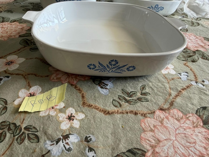 May include: A white ceramic square baking dish with rounded corners and integrated side handles. The exterior features a blue floral pattern consisting of stylized cornflowers and leaves centered on the side. The interior is smooth and white.