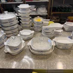 May include: A large collection of white ceramic bakeware featuring a blue floral pattern. The set includes various square and rectangular casserole dishes, some with clear glass lids, and small saucepans with handles, all arranged on a table.