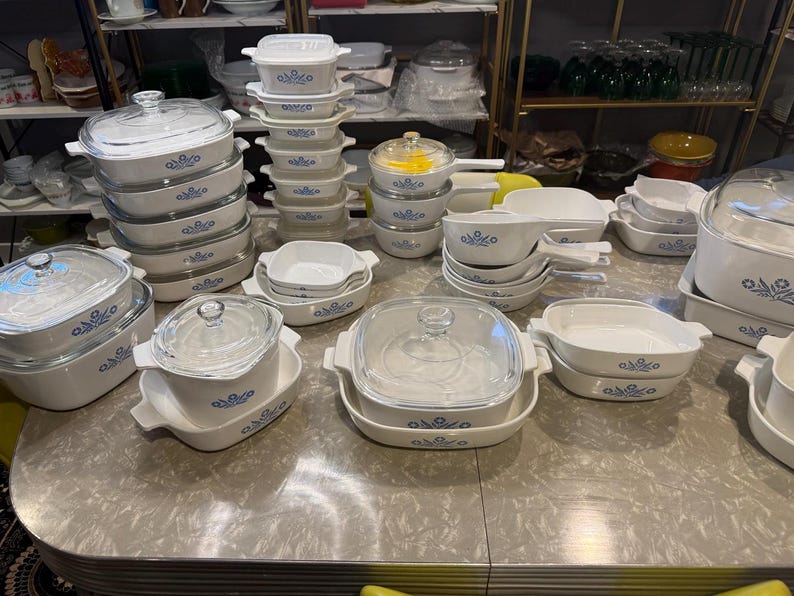 May include: A large collection of white ceramic bakeware featuring a blue floral pattern. The set includes various square and rectangular casserole dishes, some with clear glass lids, and small saucepans with handles, all arranged on a table.