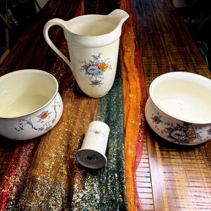 May include: A collection of antique cream-colored ceramic serving pieces with floral designs. The set includes a pitcher, two bowls with handles, and a small cylindrical container. The floral pattern features blue and orange flowers.