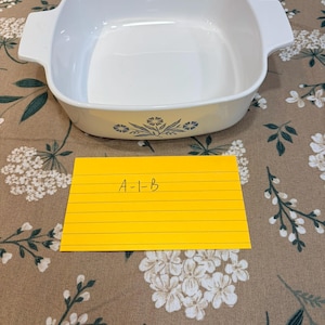 May include: A white square baking dish with integrated side handles and a blue floral pattern on the front exterior. A yellow rectangular index card is placed in front of the dish with the handwritten text A-1-B clearly visible on the paper.