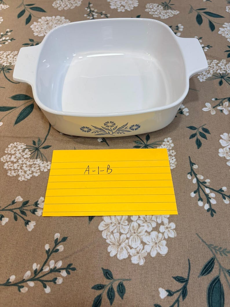 May include: A white square baking dish with integrated side handles and a blue floral pattern on the front exterior. A yellow rectangular index card is placed in front of the dish with the handwritten text A-1-B clearly visible on the paper.