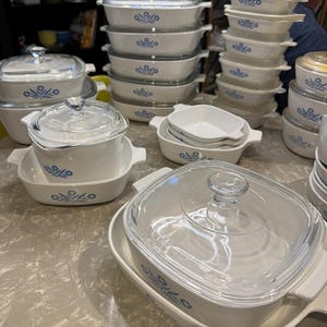 May include: A collection of white glass bakeware dishes featuring a blue floral pattern. The set includes various rectangular and square casseroles with clear glass lids, some with integrated side handles, stacked in multiple arrangements.