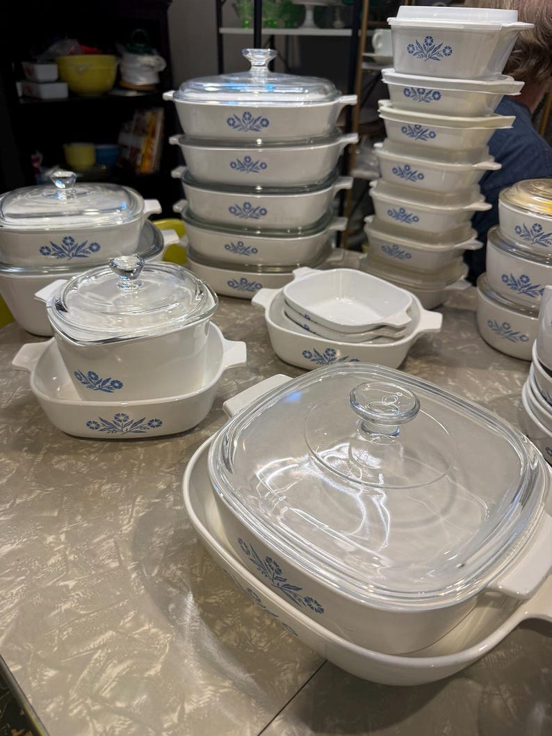 May include: A collection of white glass bakeware dishes featuring a blue floral pattern. The set includes various rectangular and square casseroles with clear glass lids, some with integrated side handles, stacked in multiple arrangements.