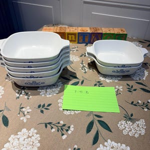 May include: A collection of white square baking dishes with integrated handles, featuring a blue floral pattern on the exterior. The set includes a stack of four dishes and a separate stack of two dishes, accompanied by a green note labeled P-41-B.