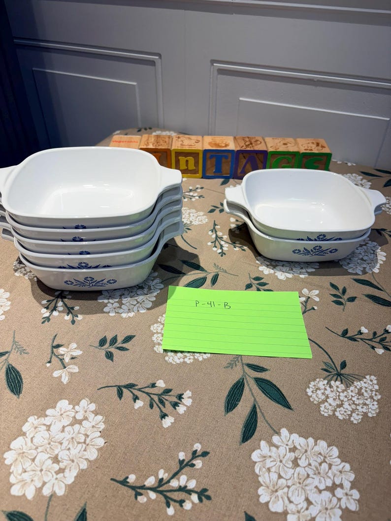 May include: A collection of white square baking dishes with integrated handles, featuring a blue floral pattern on the exterior. The set includes a stack of four dishes and a separate stack of two dishes, accompanied by a green note labeled P-41-B.