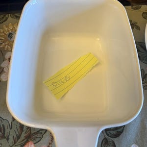 May include: A rectangular white ceramic baking dish with integrated handles on two sides. Inside the dish lies a small, torn piece of yellow lined paper with the handwritten text P-4-B clearly visible in black ink on the surface of the paper.