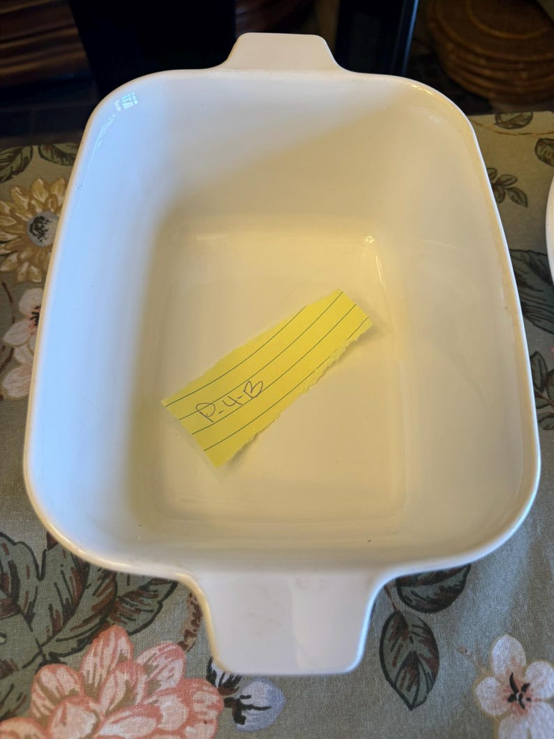 May include: A rectangular white ceramic baking dish with integrated handles on two sides. Inside the dish lies a small, torn piece of yellow lined paper with the handwritten text P-4-B clearly visible in black ink on the surface of the paper.