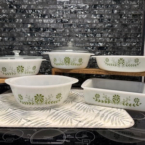 Puede incluir: Colección de fuentes Pyrex blancas vintage con un estampado floral verde. El conjunto incluye varios recipientes para hornear y una fuente rectangular. Las fuentes están dispuestas sobre una estantería de madera y una alfombrilla estampada.