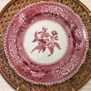 May include: A round, red and white floral patterned plate sits on a woven rattan charger. The plate features a central floral design and a decorative border. The plate's color scheme is red and white, with intricate floral patterns.