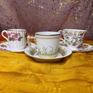 May include: Three decorative teacups and saucers are displayed on a yellow surface. The teacups feature floral and grass designs, with gold trim. The saucers complement the teacups with matching patterns. The background is a mix of pink and gold.
