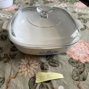 May include: A square white ceramic casserole dish with integrated side handles and a clear glass lid. The front of the dish features a blue floral pattern. A small yellow paper scrap with the handwritten text p-9-B is placed in front.