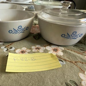 May include: Two white ceramic bowls with blue floral patterns. One bowl is open, and the other has a clear glass lid. A yellow rectangular piece of paper with the handwritten text R-82-B is placed in front of the bowls on a floral patterned surface.