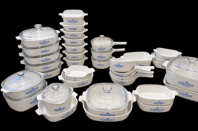May include: A large collection of white ceramic cookware featuring a blue floral pattern. The set includes various rectangular and square baking dishes, saucepans with handles, and lidded casseroles with clear glass lids, all arranged together.