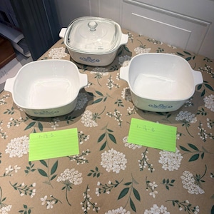 May include: Three white square ceramic casserole dishes with blue floral patterns on the sides. One dish includes a clear glass lid. Two green index cards are present, one labeled P-1 1/2-B and the other labeled P-1 3/4-B, indicating the capacity.
