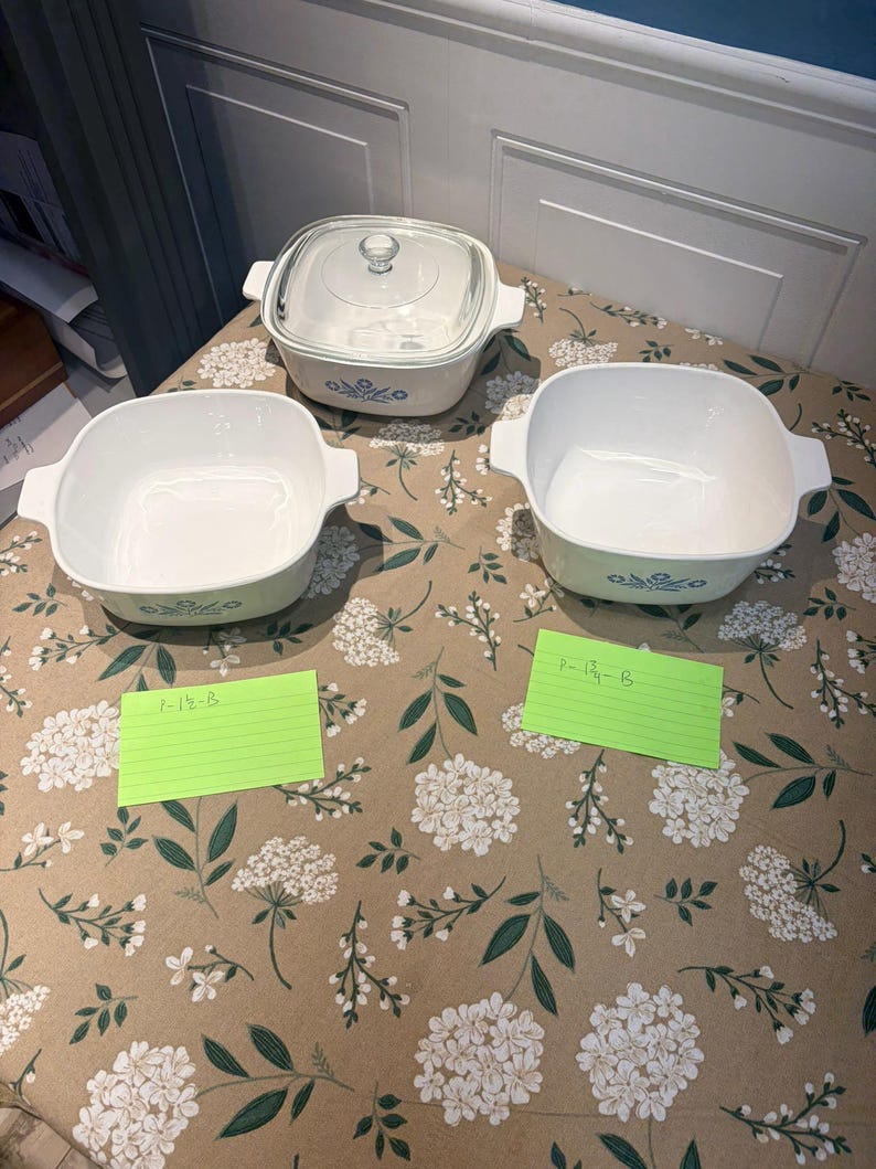 May include: Three white square ceramic casserole dishes with blue floral patterns on the sides. One dish includes a clear glass lid. Two green index cards are present, one labeled P-1 1/2-B and the other labeled P-1 3/4-B, indicating the capacity.