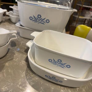 May include: A collection of white ceramic bakeware dishes featuring a blue floral pattern. The set includes a large rectangular casserole dish with a clear glass lid, a square baking dish, and an oval serving dish, all with integrated handles.