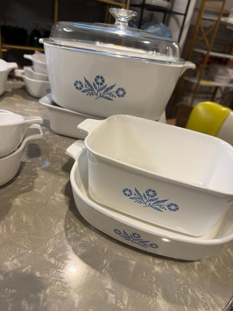 May include: A collection of white ceramic bakeware dishes featuring a blue floral pattern. The set includes a large rectangular casserole dish with a clear glass lid, a square baking dish, and an oval serving dish, all with integrated handles.