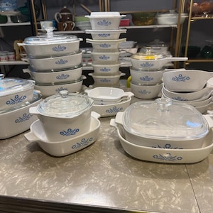 May include: A large collection of white ceramic bakeware featuring a blue floral pattern. The set includes various rectangular and square casserole dishes, some with clear glass lids, and saucepans with handles, all arranged on a table.