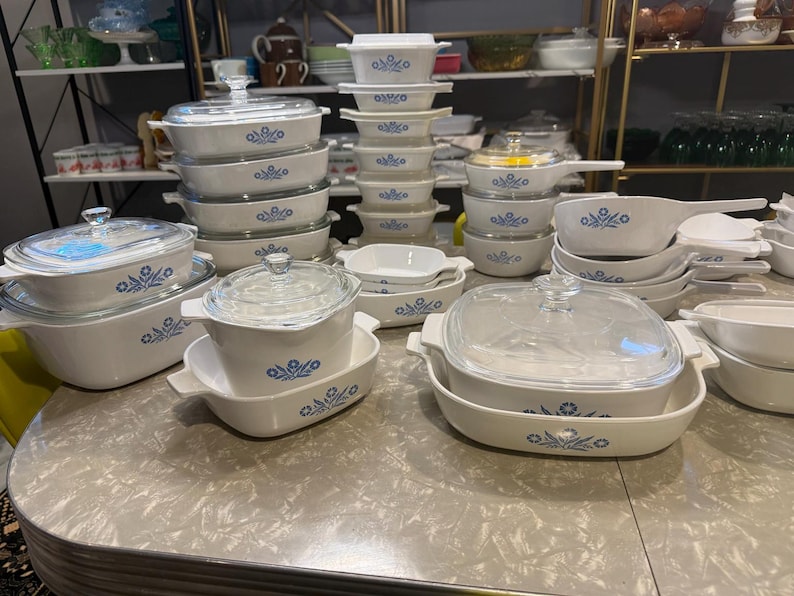 May include: A large collection of white ceramic bakeware featuring a blue floral pattern. The set includes various rectangular and square casserole dishes, some with clear glass lids, and saucepans with handles, all arranged on a table.
