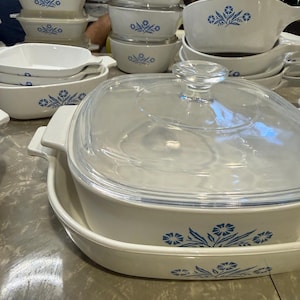 May include: A collection of white ceramic cookware featuring a blue floral pattern. The set includes various square and rectangular baking dishes, some with clear glass lids, and saucepans with handles, all decorated with a consistent blue flower motif.