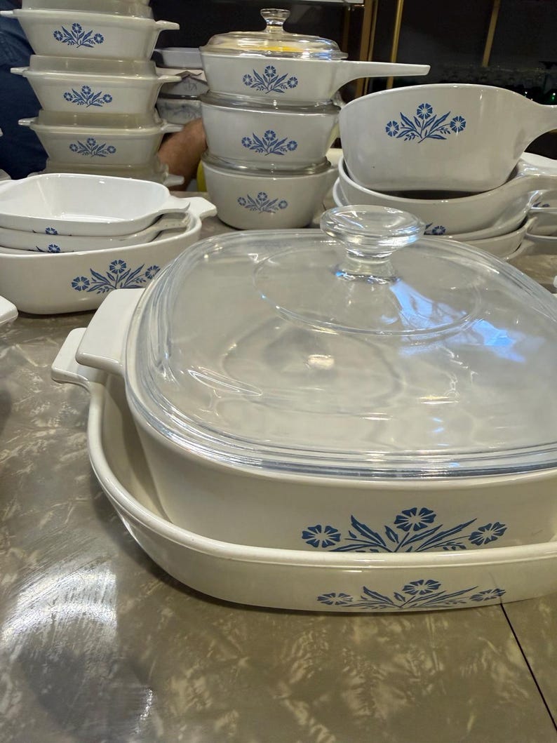May include: A collection of white ceramic cookware featuring a blue floral pattern. The set includes various square and rectangular baking dishes, some with clear glass lids, and saucepans with handles, all decorated with a consistent blue flower motif.