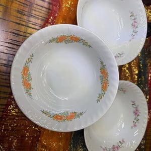 May include: Three white ceramic bowls with floral designs. Two bowls have orange and green floral patterns, while the third has pink and green floral patterns. The bowls are arranged on a patterned surface.