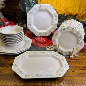 Può includere: Una collezione di stoviglie ottagonali bianche con un motivo floreale in colori pastello. Il set comprende piatti, ciotole e un vassoio da portata. I piatti sono impilati ed esposti su una superficie di legno.