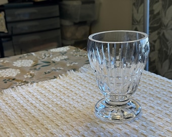 May include: A clear, cut-glass goblet with a wide, fluted bowl and a round base. The glass has a textured, vertical pattern. The goblet sits on a white, textured tablecloth. The background includes a floral patterned tablecloth and a dark cabinet.
