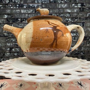May include: A handcrafted ceramic teapot with a lid and handle. The teapot features a brown and tan speckled design with a silhouette of a figure. It sits on a white, decorative, tiered stand.